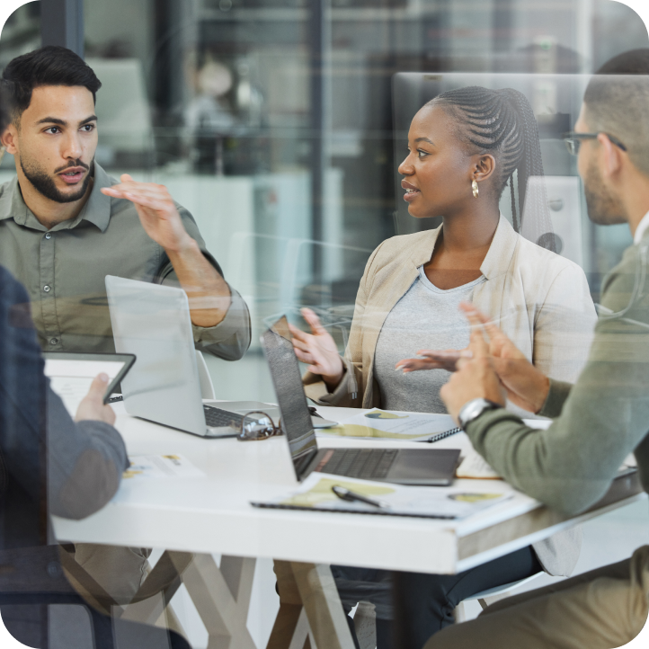 A business meeting is happening through window glass with three individuals. Laptops and pens and paper sit on the table.