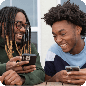 Two young men sitting at a table, smiling while looking at their smartphones.