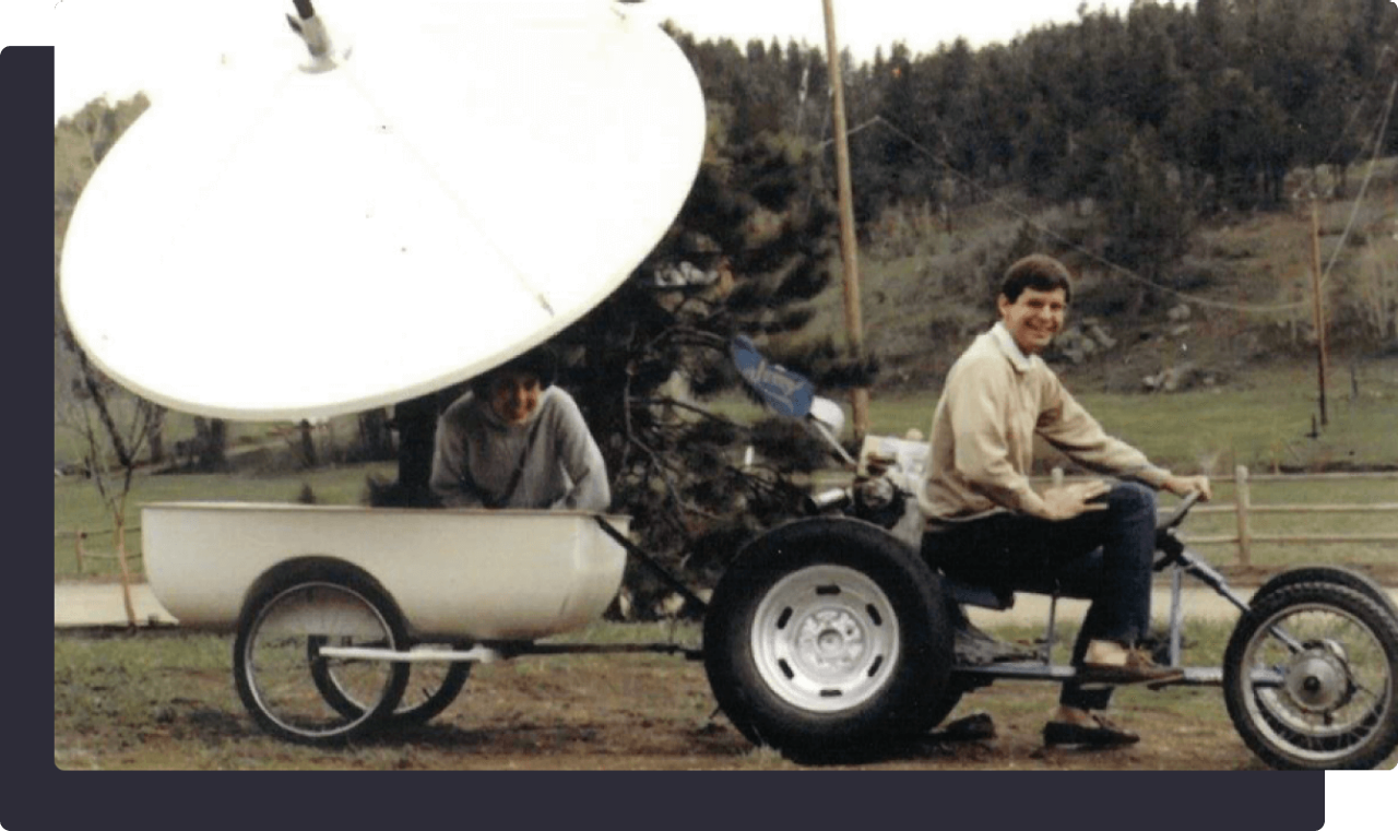 A man riding a custom-built tricycle towing a satellite dish with another person sitting inside.