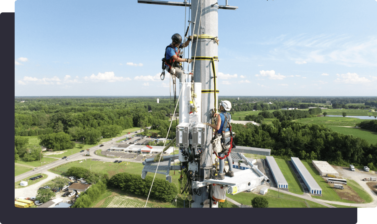 Two workers in safety gear climbing a communications tower against a rural landscape.
